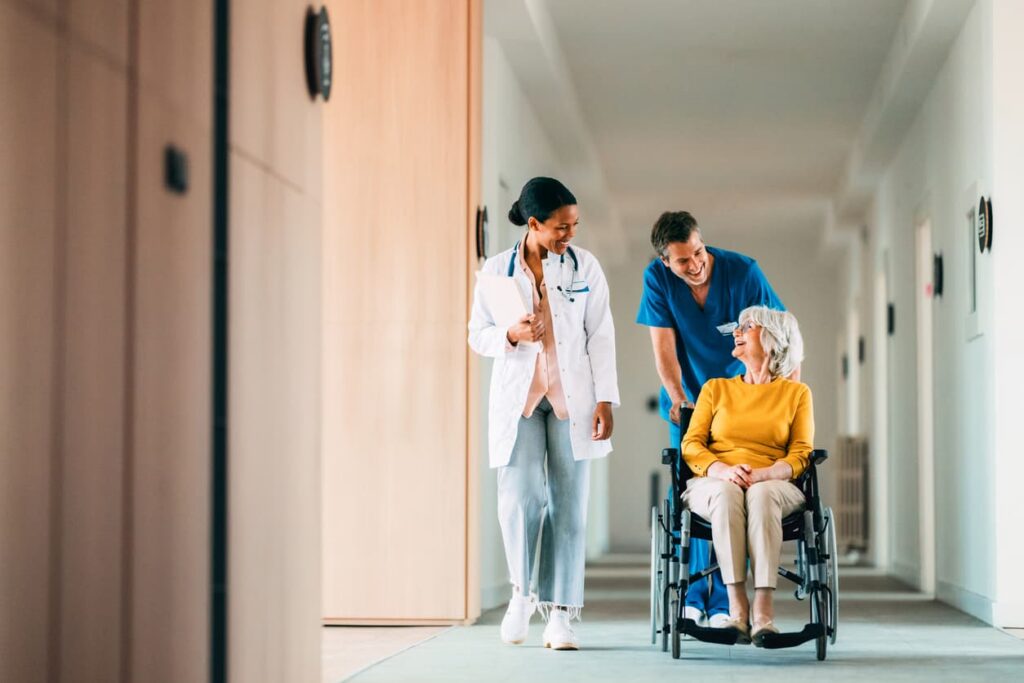 Doctor and nurse assisting elderly woman in wheelchair in hospital hallway, representing senior medical care and nursing home safety.